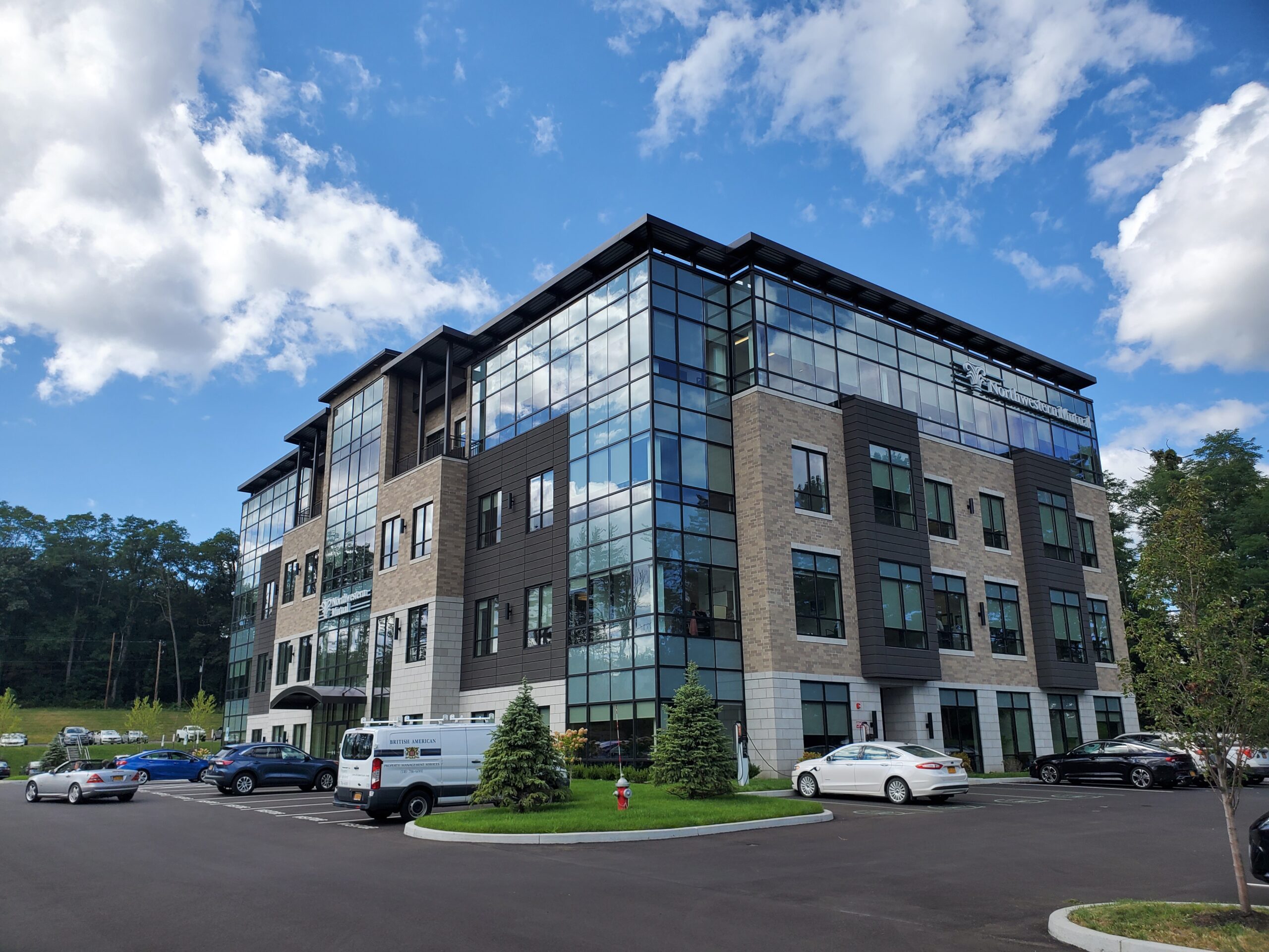Photo of the exterior of Northwestern Mutual's headquarters in Latham, New York. Taken on a sunny day, blue sky with white clouds set behind building, greenery and a parking lot with a few cars are also in the image. Building features materials in earthtones as well as many windows reflecting the sky.