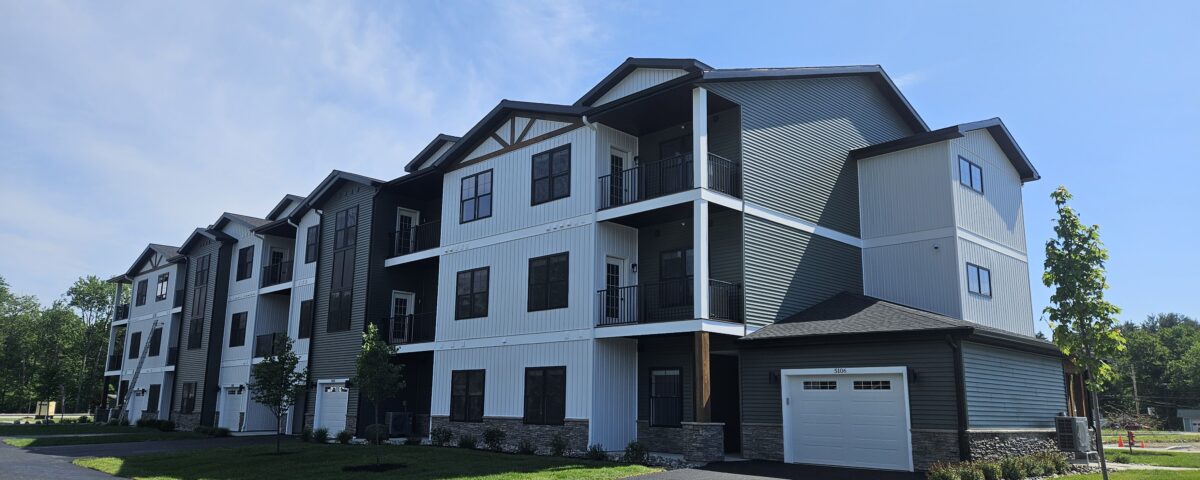 Photo of a three story apartment building with black windows, a variety of surface materials and colors, garages, and covered decks. Building is set against a blue sky with greenery surrounding.