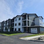 Photo of a three story apartment building with black windows, a variety of surface materials and colors, garages, and covered decks. Building is set against a blue sky with greenery surrounding.