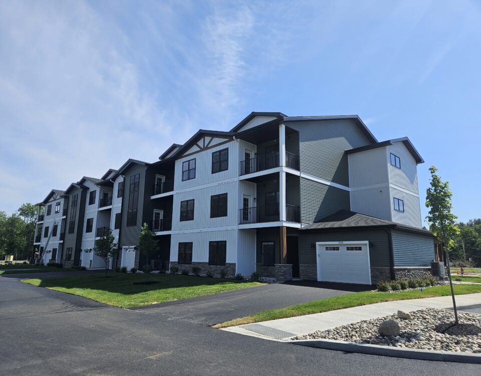 Photo of a three story apartment building with black windows, a variety of surface materials and colors, garages, and covered decks. Building is set against a blue sky with greenery surrounding.