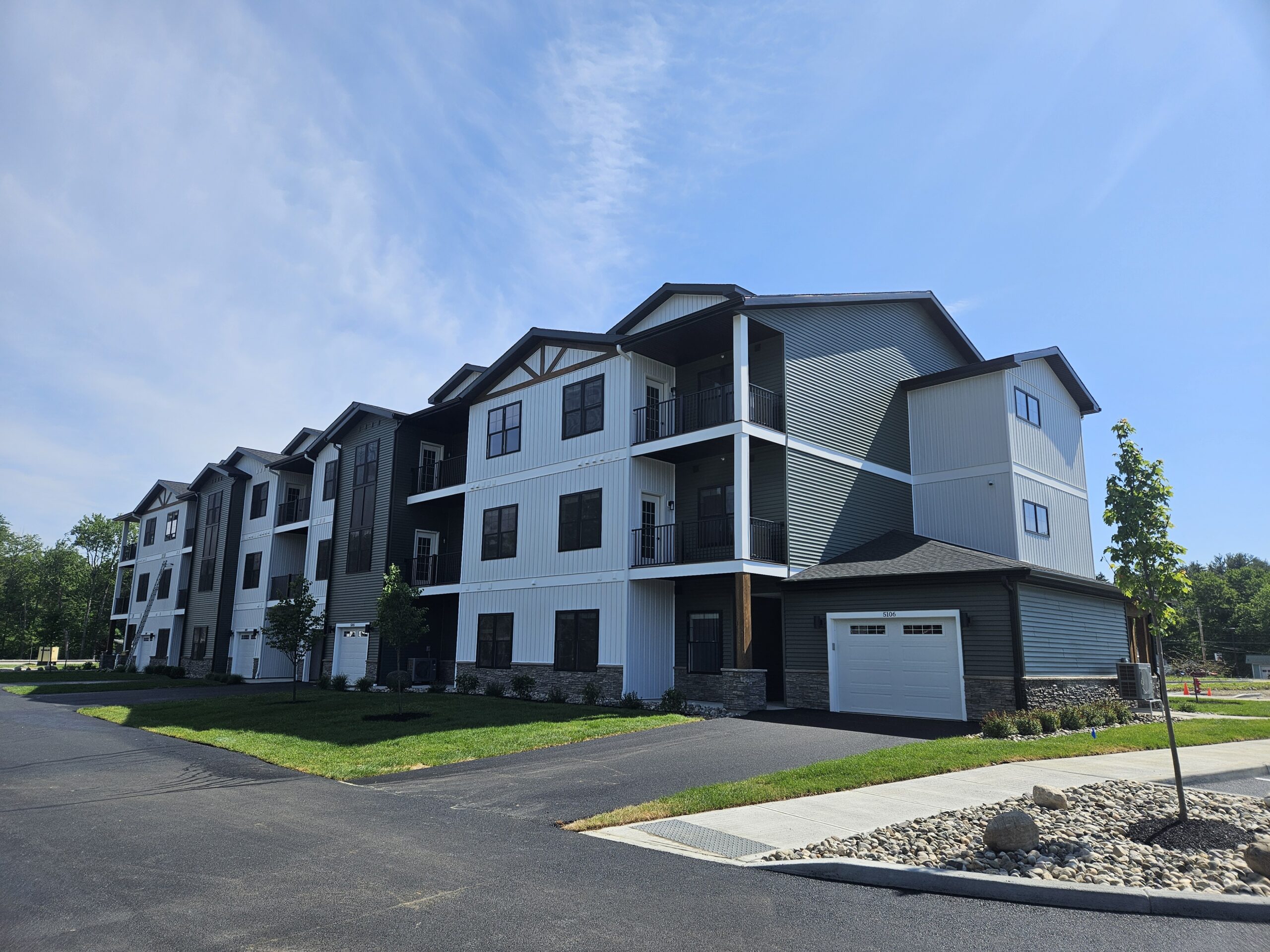 Photo of a three story apartment building with black windows, a variety of surface materials and colors, garages, and covered decks. Building is set against a blue sky with greenery surrounding.